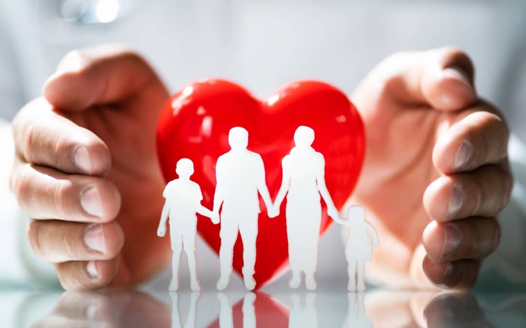 a small family made of paper standing in front of a red heart with a man's hands cupping them loosely