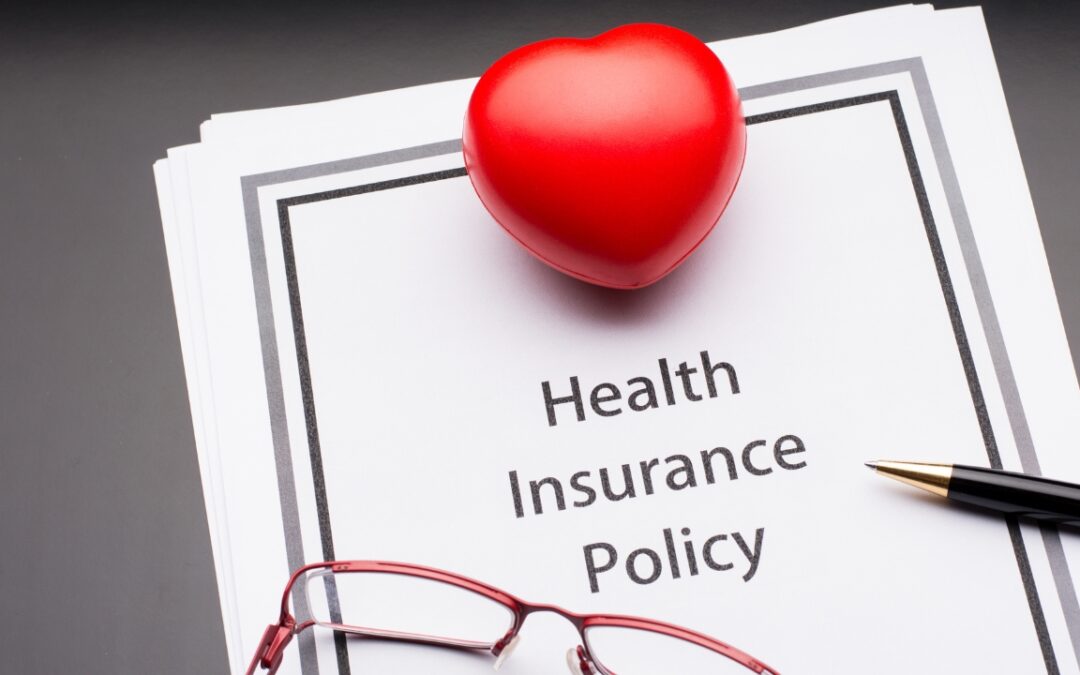 a piece of paper with the words "health insurance policy" printed on it; there is a red pair of glasses and a small red heart sitting atop the paper