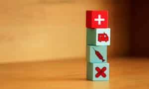 a stack of wooden blocks; the top one has a small white plus sign, the next has a little ambulance, the next has a syringe, and the last one has crossed bandages