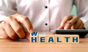 a person flipping the first of a line of wooden blocks that spell "health," except the "h" is turned into a "w" changing the word from "health" to "wealth"