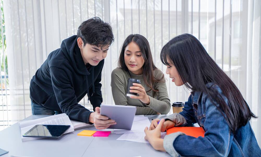 a group of three students looking at a tablet or laptop in the middle of a table littered with class papers and notes